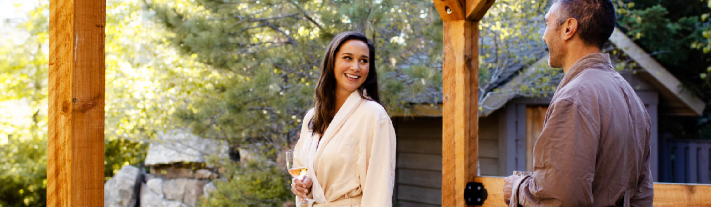 A woman holding a drink smiles at a man outdoors near a wooden structure at Suncadia Resort. Trees and a building provide a serene backdrop, capturing the relaxed ambiance of this Washington getaway.