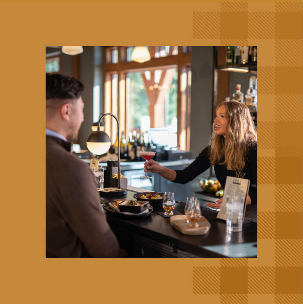 At a bustling Cle Elum restaurant, a woman serves a drink to a man at a bar counter. Plates of food and glasses adorn the counter, while the background reveals shelves of bottles and large windows that let in the stunning views reminiscent of Suncadia Resort's enchanting ambiance.