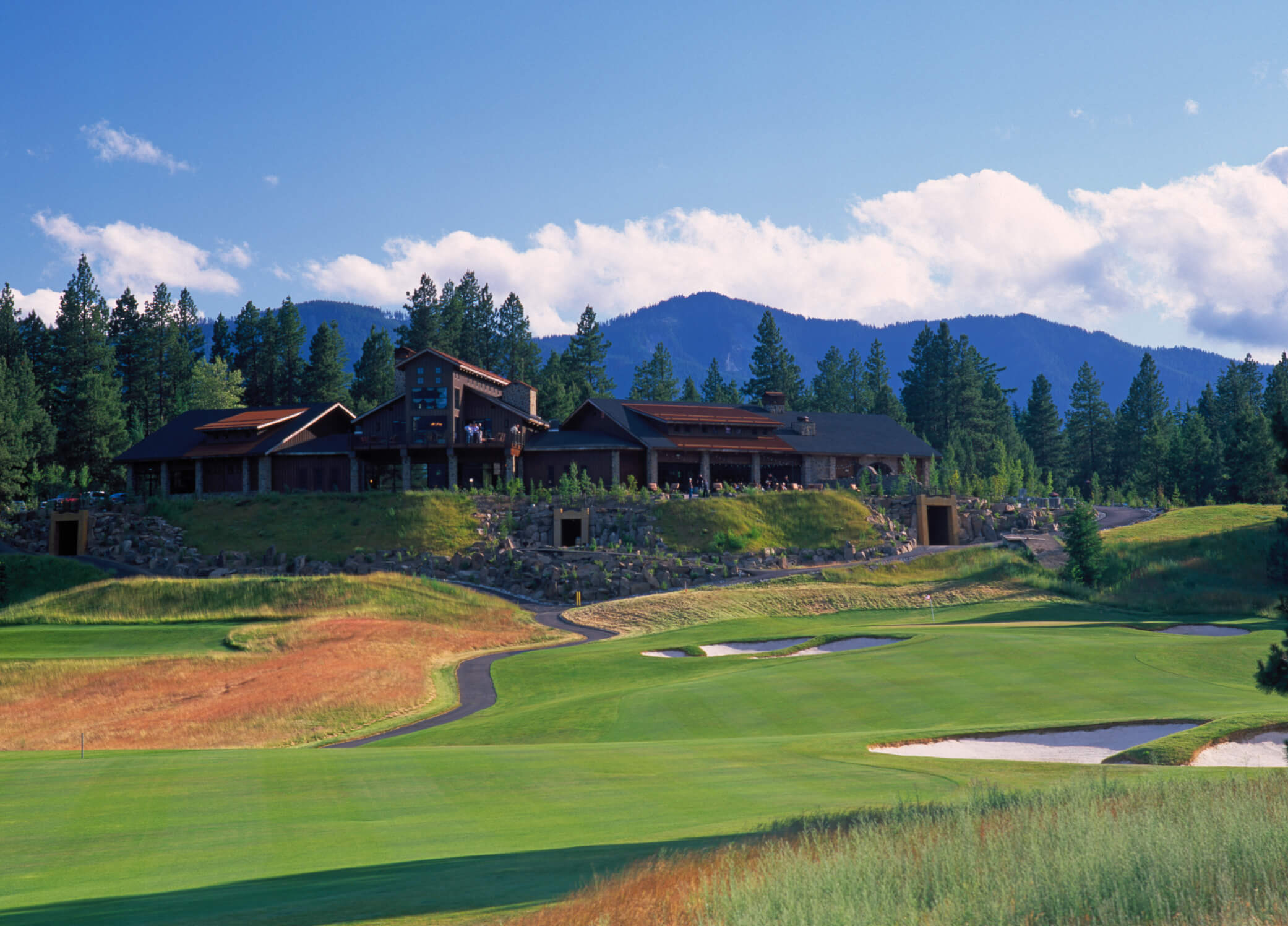 The golf course landscape at Suncadia Resort features a charming clubhouse surrounded by lush fairways, sand traps, and towering pines under a partly cloudy sky, set against the majestic backdrop of Washington's distant mountains.