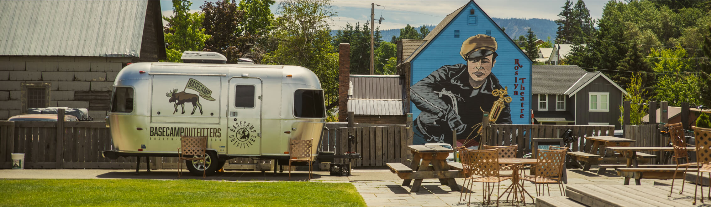 A vintage trailer and outdoor seating sit in front of a mural depicting a person holding a tool, evoking the rustic charm reminiscent of Suncadia Resort.