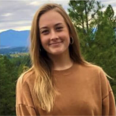 A young woman with long hair stands outdoors, donning a brown sweater against the stunning backdrop of trees and mountains at Suncadia Resort Washington.