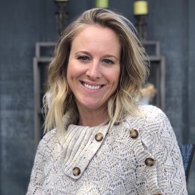 A person with shoulder-length blond hair smiling, wearing a light-colored, buttoned sweater in front of the charming backdrop of Suncadia.