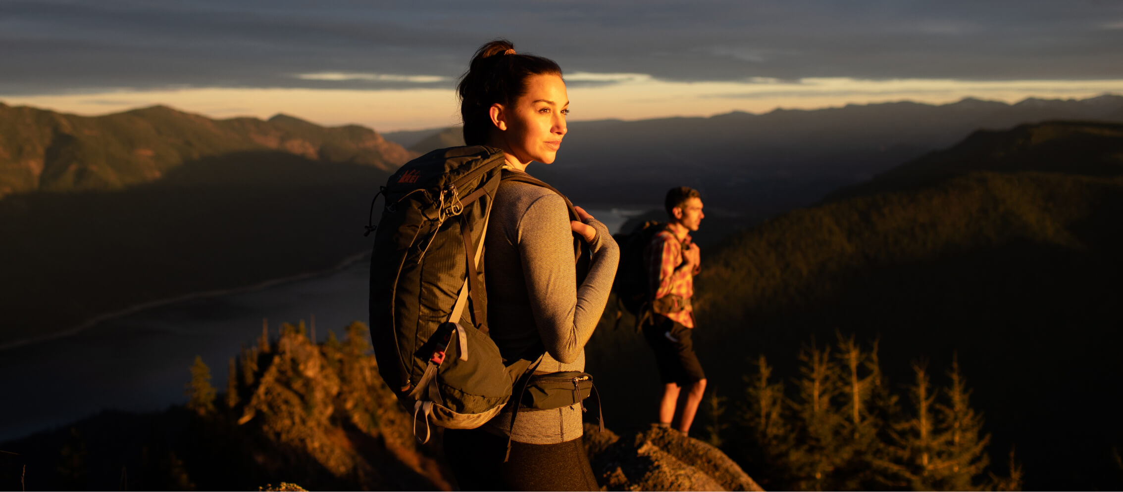 Two hikers with backpacks stand on a mountain ledge, overlooking a valley at sunset near the Suncadia Resort in Washington.