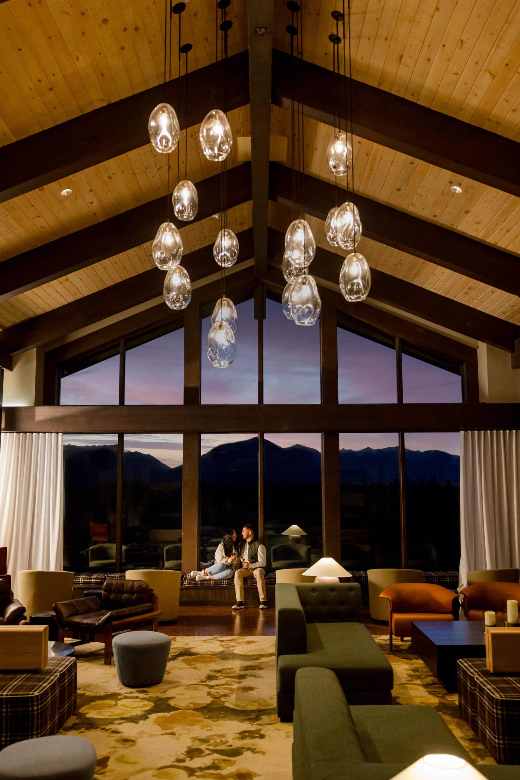 A spacious lodge interior at Suncadia Resort features a high wooden ceiling, modern seating, and a large window with a stunning mountain view. As the evening light filters in, two people sit near the window, reminiscent of cozy evenings at Cle Elum restaurants.