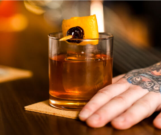 A tattooed hand rests next to a glass filled with a rich brown liquid, garnished with an orange slice and a cherry on a toothpick. The drink exudes the warmth and charm reminiscent of Suncadia homes, while the glass sits elegantly on a square coaster.