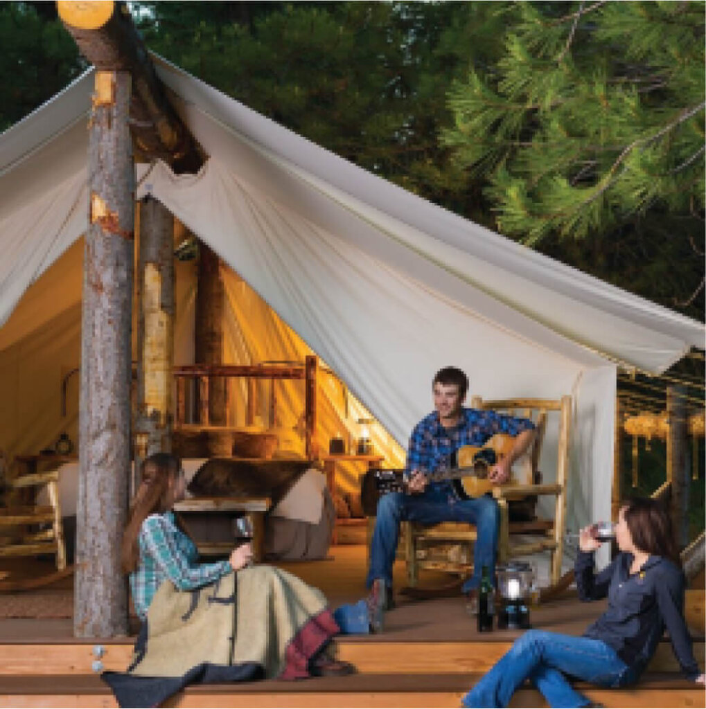 Three friends relax outside a softly lit canvas tent nestled in the forest, enjoying wine and guitar melodies. With chairs and a blanket on the wooden deck, it feels like an evening at home near Suncadia real estate's serene beauty.