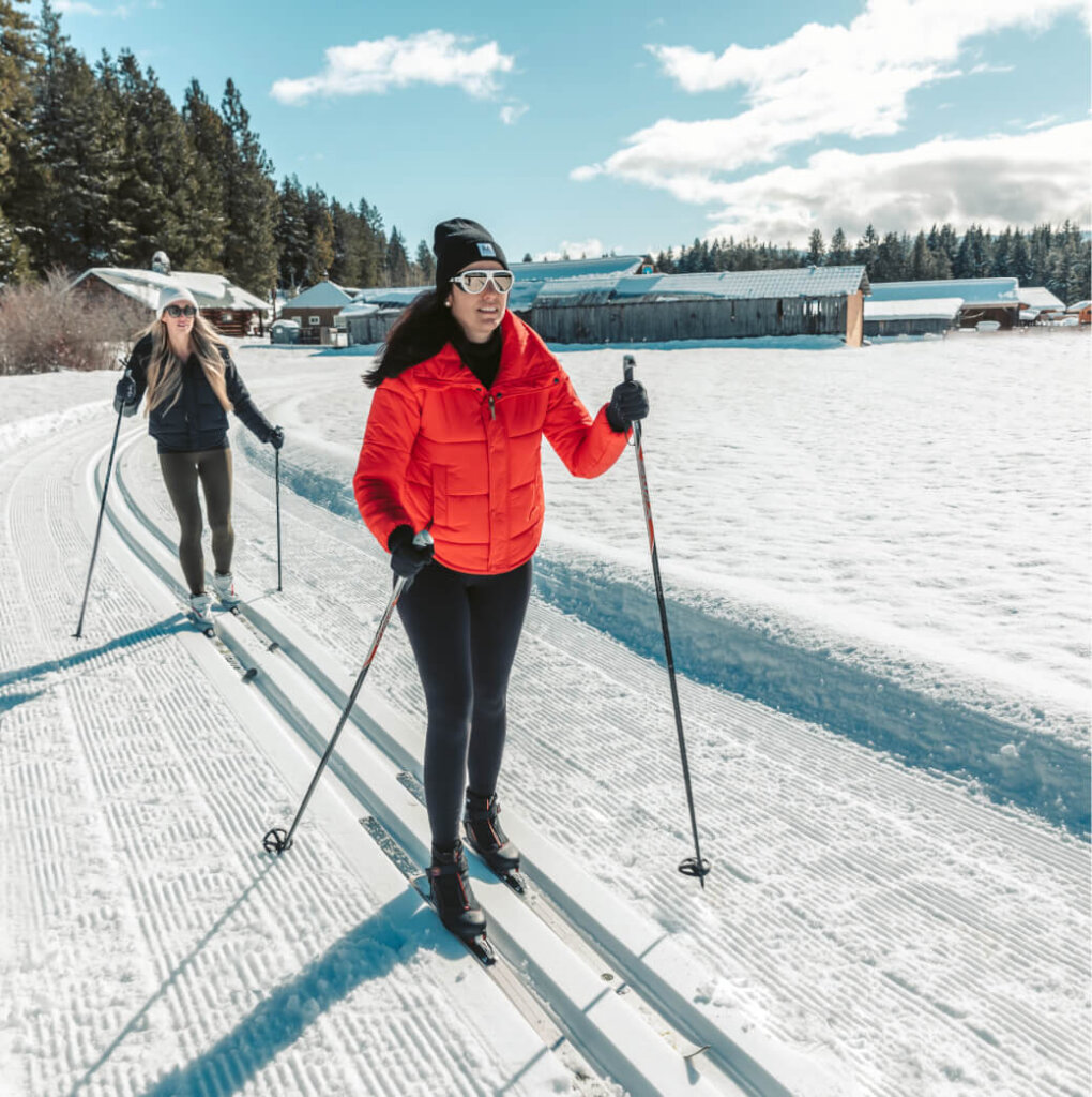 Two people cross-country skiing on a snowy trail, wearing winter gear, with the scenic backdrop of Suncadia Resort Washington. A wooden building and tree line stand under a partly cloudy sky, evoking the charm of nearby Cle Elum restaurants inviting explorers to unwind after their adventure.