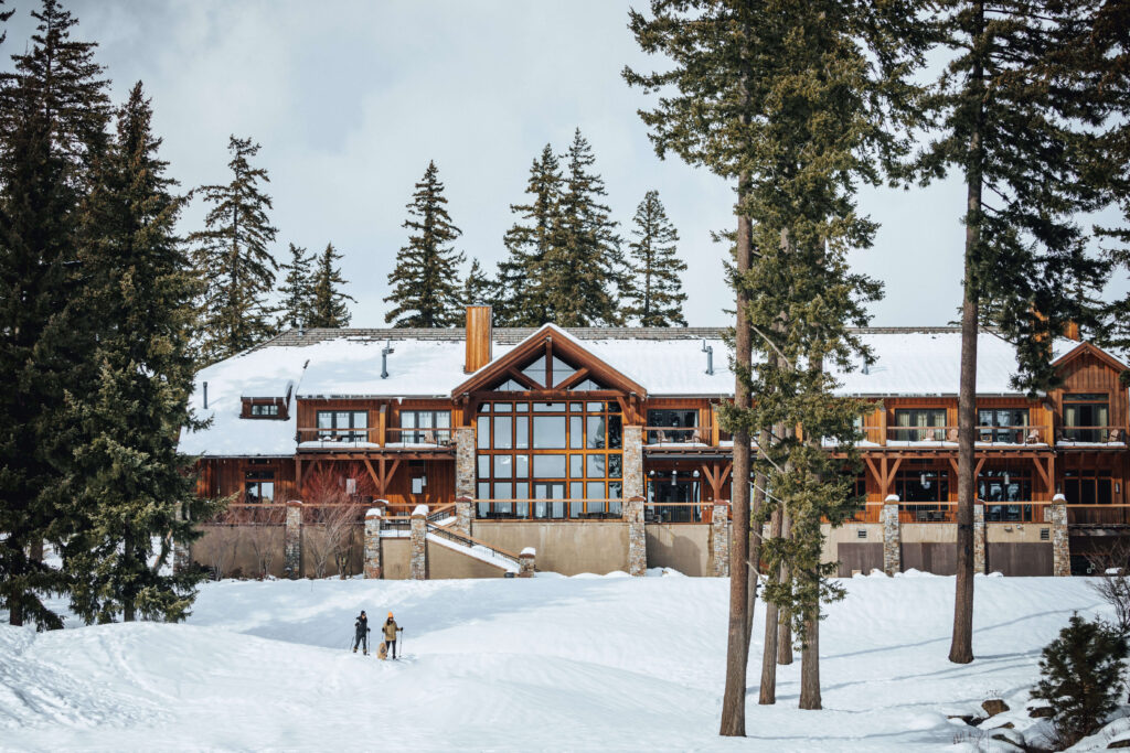 A large, rustic lodge stands in a snowy landscape surrounded by tall pine trees at Suncadia Resort, with two people walking in the snow in the foreground.