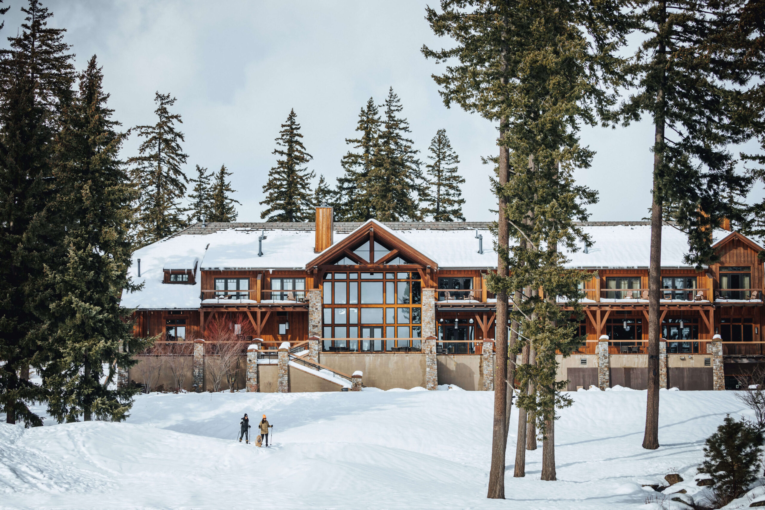 A large, rustic lodge stands in a snowy landscape surrounded by tall pine trees at Suncadia Resort, with two people walking in the snow in the foreground.