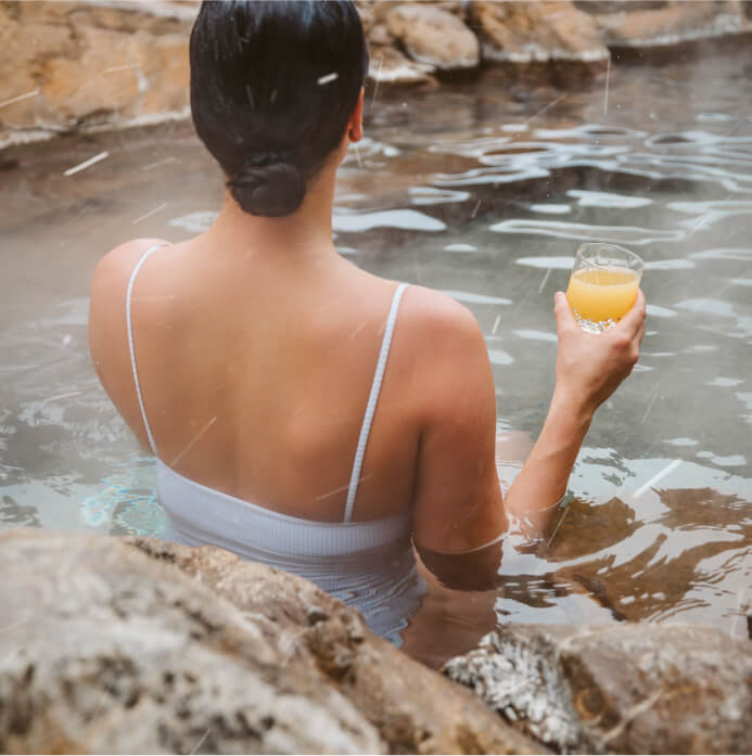 At Suncadia Resort, a person in a swimsuit relaxes in a natural hot spring, savoring a glass of orange juice. Rocks and steam create the perfect serene backdrop for this Washington escape.