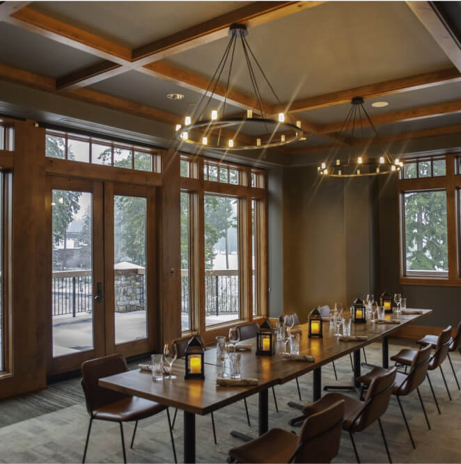 A dining room in a Suncadia Resort home features a long table, brown chairs, and modern chandeliers. Large windows overlook the scenic outdoor area, and the table is elegantly set with glassware and lanterns. Perfect for embracing both comfort and style in your Suncadia real estate dream.