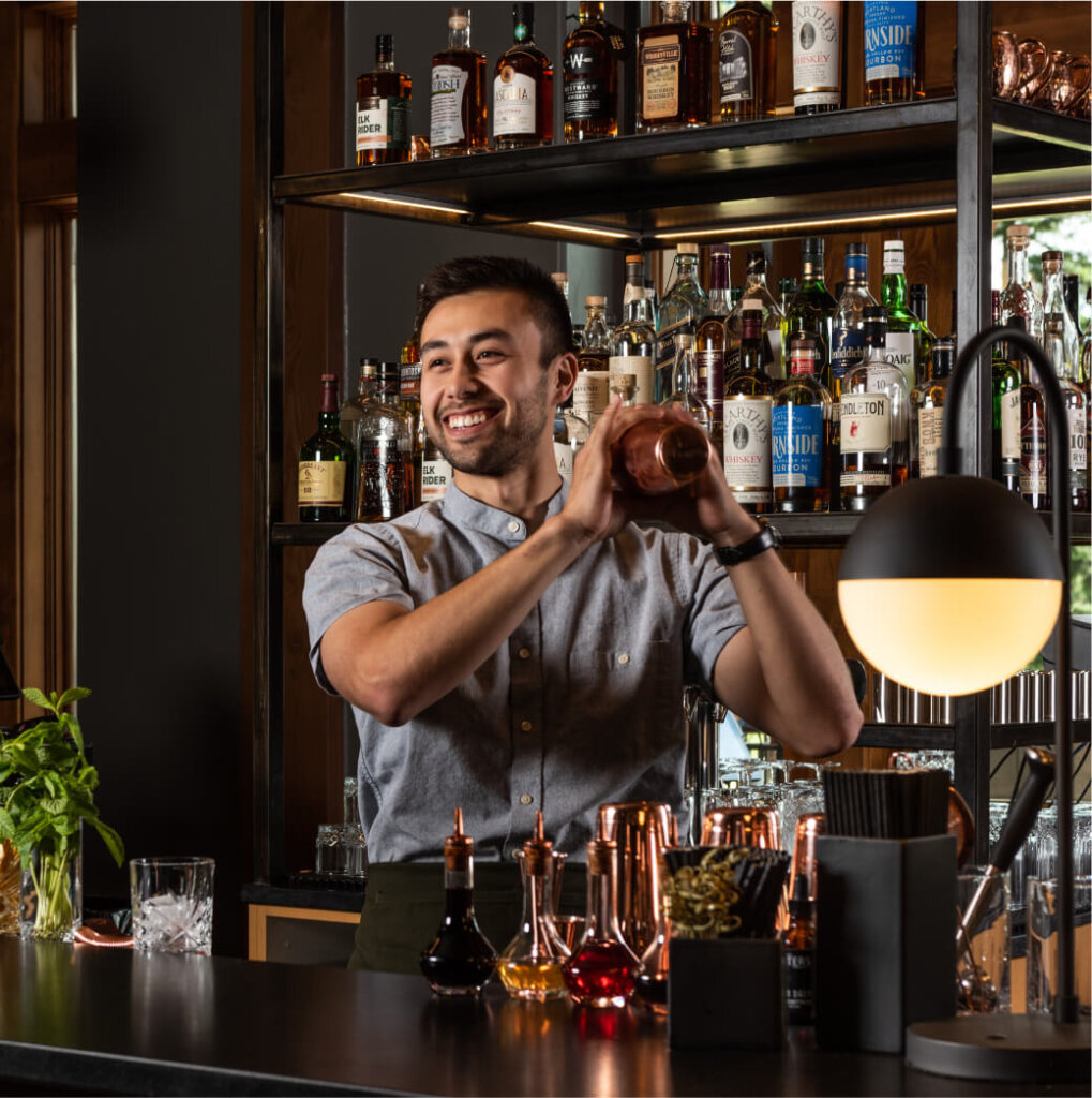 The bartender at Suncadia Resort smiles warmly while skillfully shaking a cocktail shaker behind a bar brimming with various bottles and garnishes.