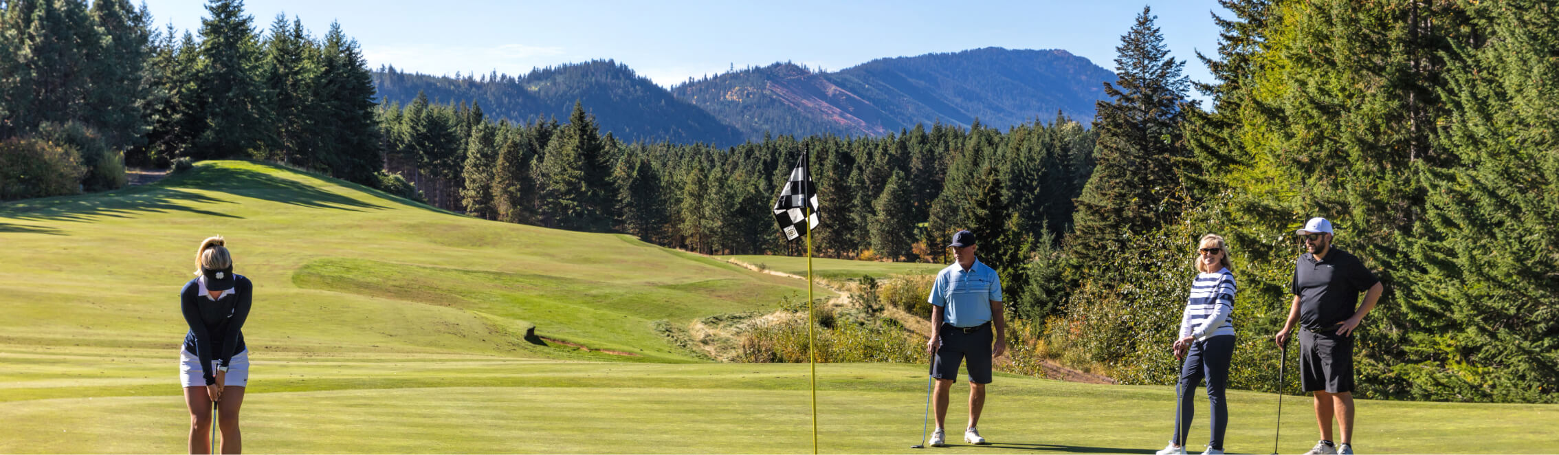 Four people stand on a stunning golf course at Suncadia, with majestic mountains and lush trees in the background. One person is focused on putting as the others watch in anticipation, taking in the serene beauty that makes Suncadia homes and real estate so desirable.