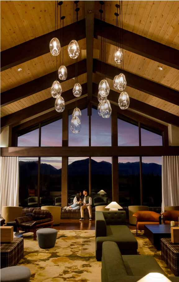 A spacious lounge at Suncadia Resort features modern furniture and a high wooden ceiling. Large windows present a stunning mountainous sunset view. Two people sit in the center, beneath artistic hanging lights, enjoying the serene elegance synonymous with Suncadia homes.