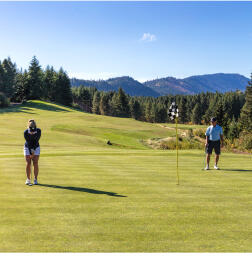 Two people enjoy a game of golf on the lush Suncadia Resort course. One is about to putt while the other watches, framed by trees and mountains under a clear blue sky, just a short drive from Cle Elum restaurants for the perfect après-golf dining experience.