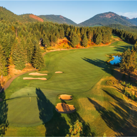 Aerial view of a golf course at Suncadia Resort, surrounded by trees with sand traps, a pond, and mountains in the background.