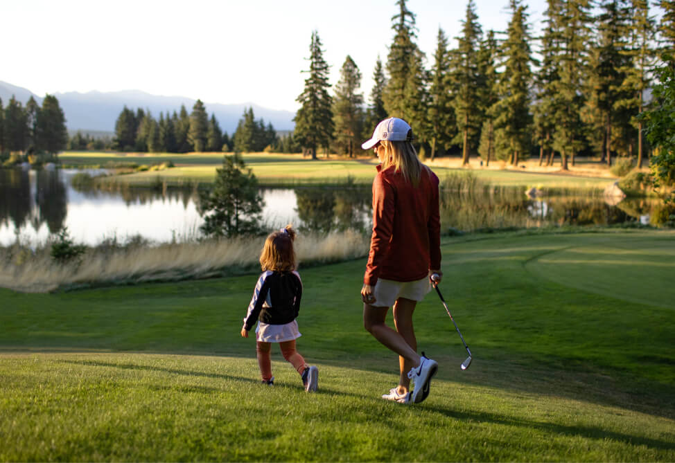 A woman and a child stroll on the scenic golf course at Suncadia Resort, near a tranquil lake, embraced by trees and mountains. The woman holds a golf club gracefully.