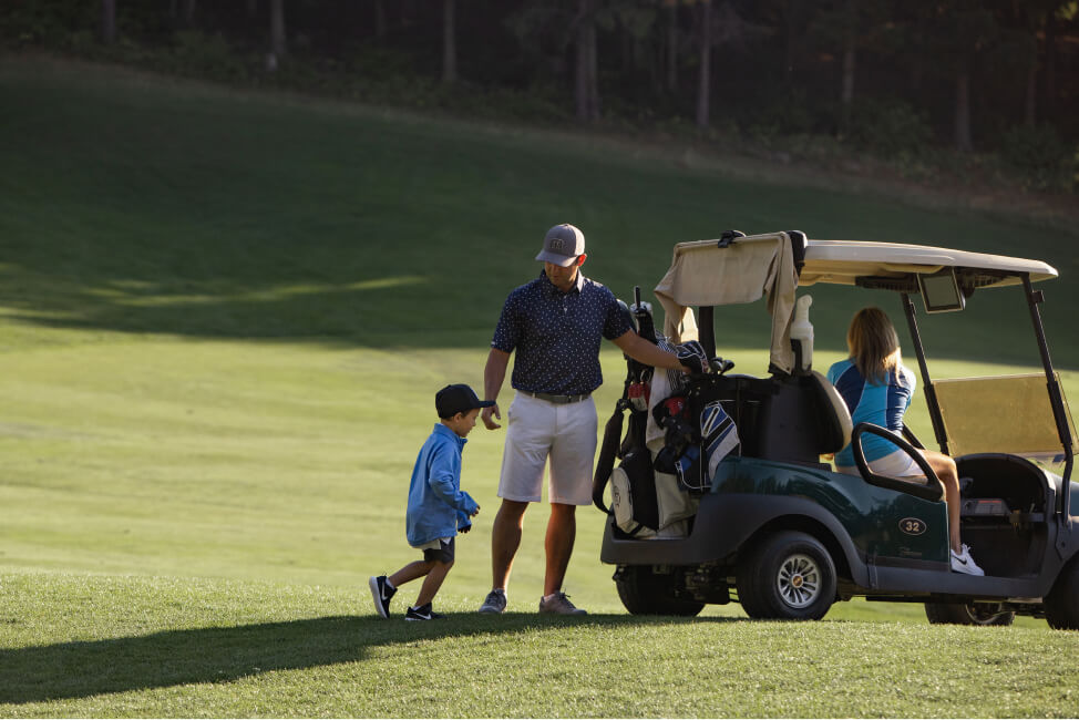 A man and child stand near a golf cart with golf bags, while a woman relaxes inside. They are enjoying the lush greens of Suncadia Resort on a sunny day, a perfect setting for exploring the beauty of Suncadia real estate.