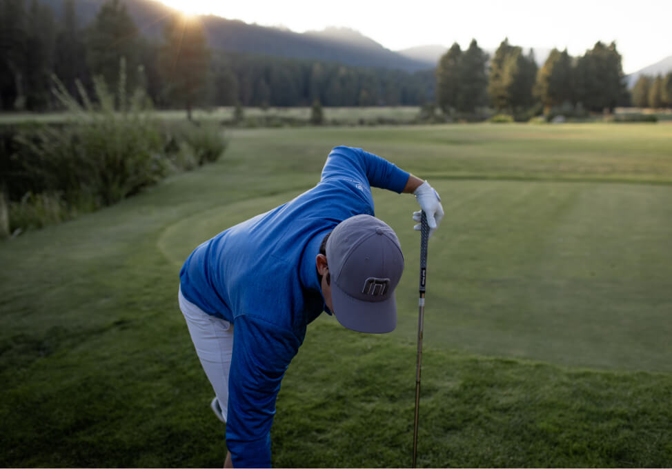 A golfer in a blue jacket and cap bends down to pick up a ball on the dewy grass at Suncadia Resort, as the sunrise casts a golden glow over the course.