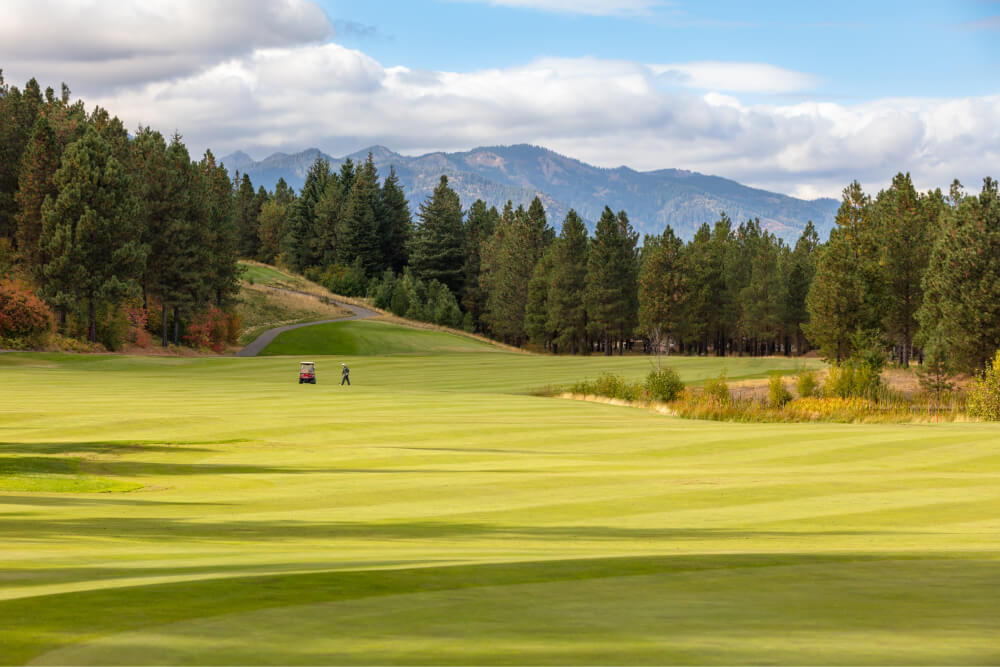 A person strolls through a golf course in Suncadia, surrounded by trees and mountains under a cloudy sky.