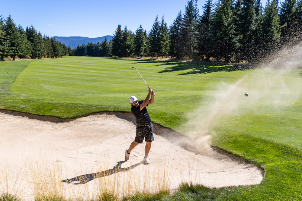 Amid the towering trees at Suncadia Resort, a golfer expertly hits a ball out of a sand trap under the clear, sunny sky.