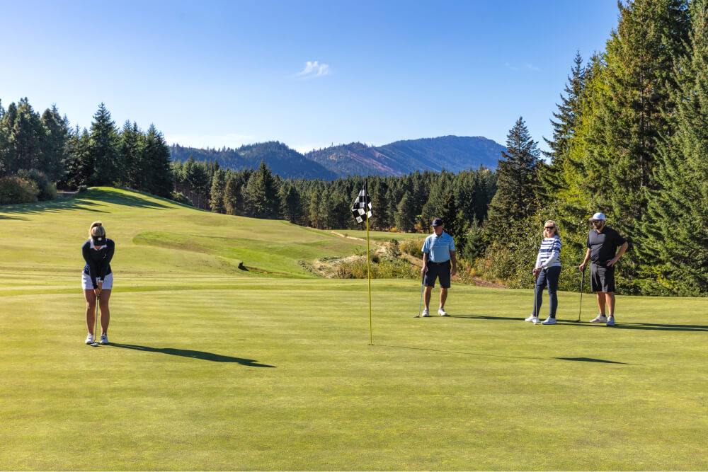 A person prepares to putt on a golf green at Suncadia Resort as three others watch. Trees and mountains create a stunning backdrop under a clear sky.