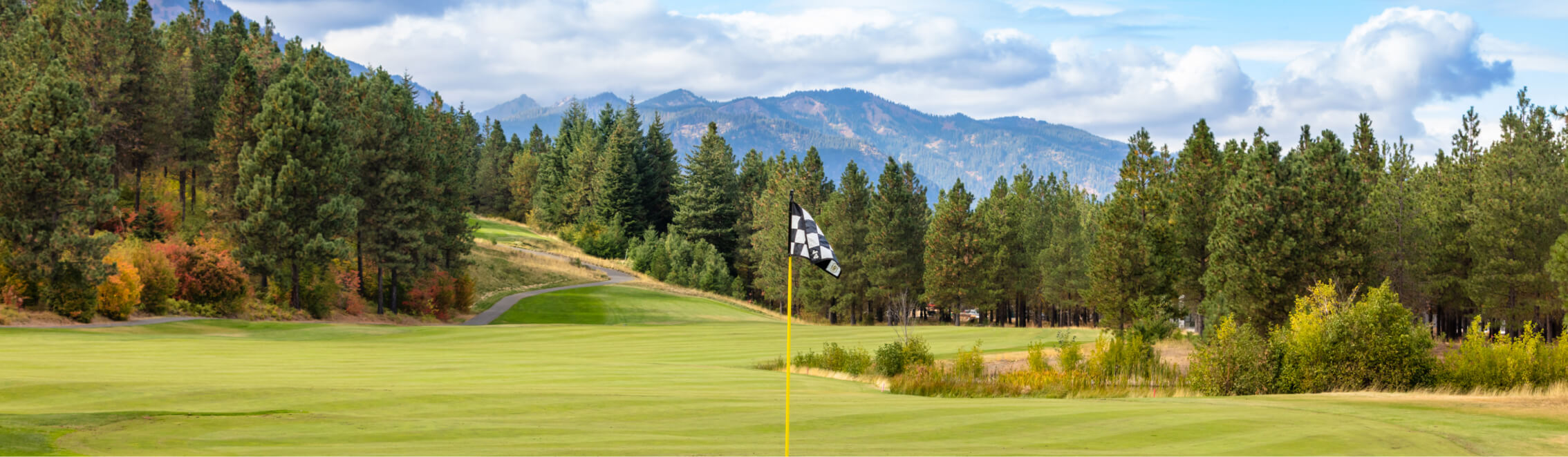 Nestled within the scenic beauty of Suncadia Resort Washington, this golf course features a checkered flag on the green, surrounded by lush trees and majestic mountains under a partly cloudy sky—a perfect backdrop for those exploring Suncadia real estate opportunities.