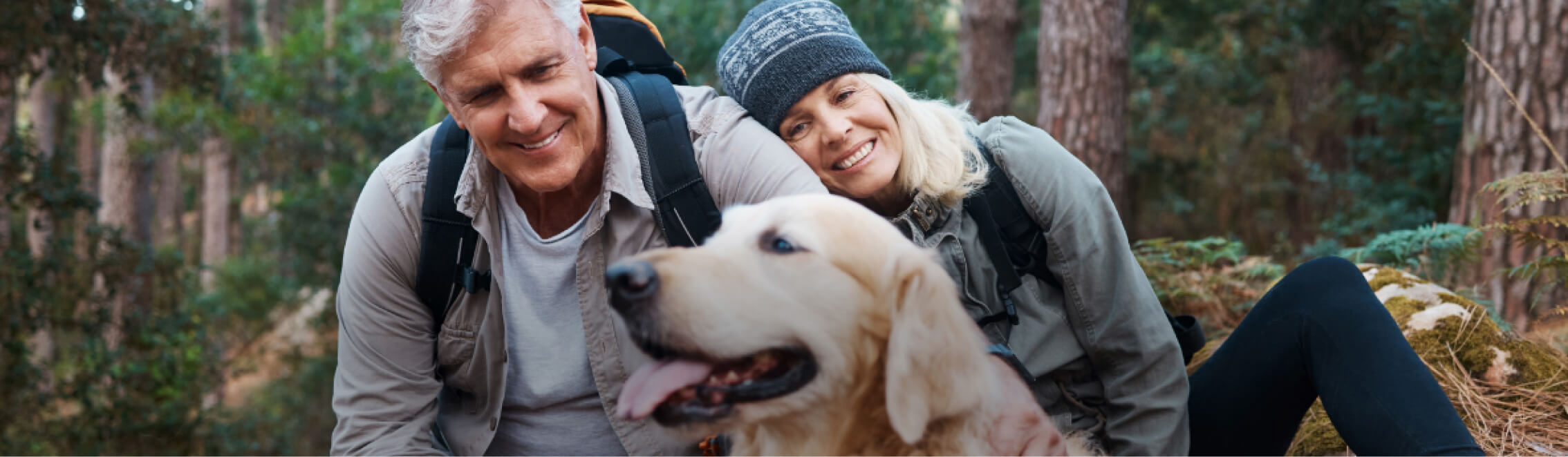 Two people in outdoor attire are sitting with a golden retriever in a serene forest setting near Suncadia Resort, Washington. Both are smiling and wearing backpacks, perhaps taking a break before exploring the nearby Cle Elum restaurants.