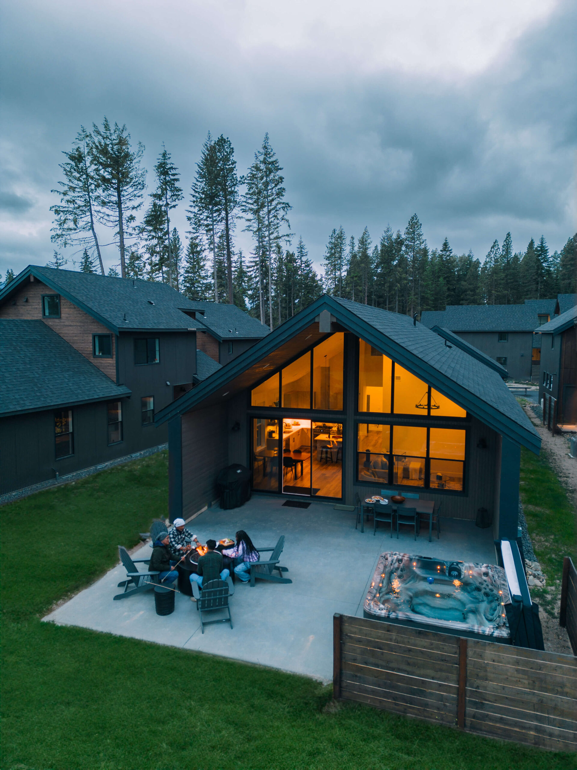 A group of people sit around a fire pit on a patio next to a modern Suncadia house with large windows. Nearby, a hot tub is filled with water. The scene is set in a wooded area at dusk, offering the perfect blend of nature and luxury that Suncadia real estate uniquely provides.