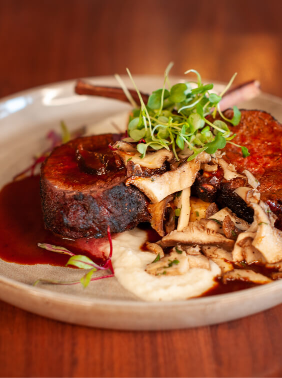 Experience a gourmet dish reminiscent of Cle Elum restaurants, featuring a succulent piece of meat on puree, topped with mushrooms, microgreens, and a savory sauce, all elegantly presented on a ceramic plate.