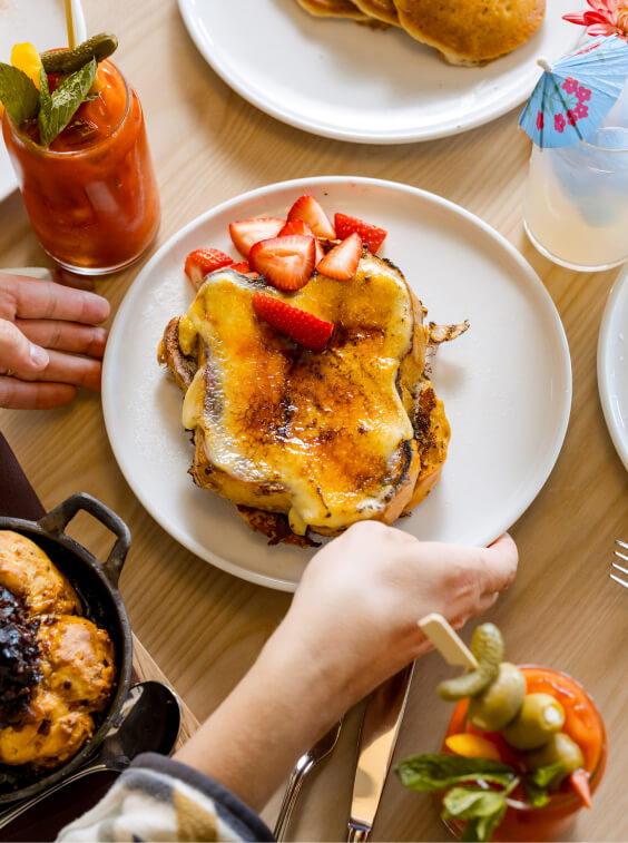 Two people reaching for a plate of French toast topped with strawberries at Suncadia Resort. Nearby are a Bloody Mary and a tropical drink, adding to the relaxing atmosphere of Washington's scenic getaway.