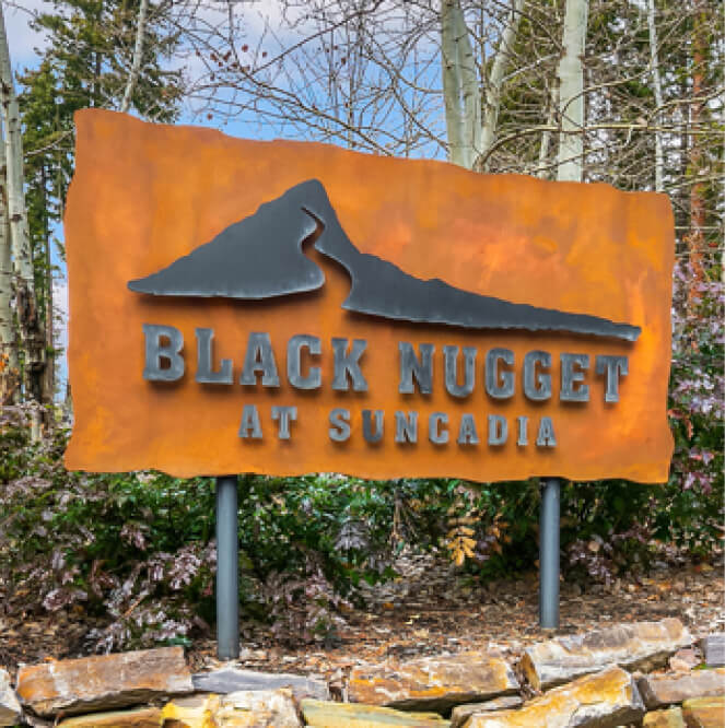 Rustic metal sign reading "Black Nugget at Suncadia" with a mountain silhouette, set among trees and rocks, inviting you to explore the nearby charm of Cle Elum restaurants.