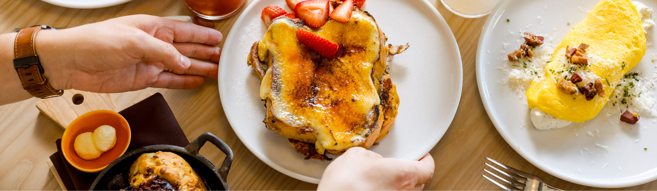 A person serves French toast topped with strawberries, next to an omelette and a small skillet with butter and syrup, capturing the cozy dining vibe reminiscent of mornings at Suncadia Resort in Washington.