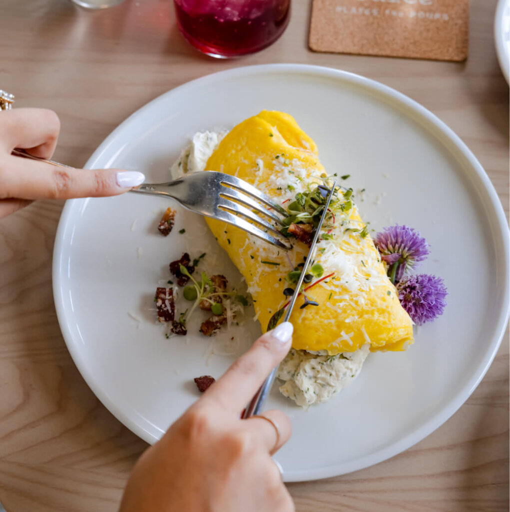 At Suncadia Resort, a person cuts into a yellow omelet garnished with herbs and purple flowers on a pristine white plate.