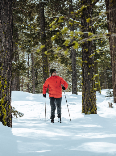 A person in a red jacket is snowshoeing through a snowy forest with tall trees, capturing the serene beauty of Suncadia Resort Washington as they explore the winter wonderland.