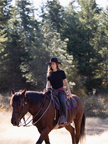 Under the bright sun, a person in a black hat rides a brown horse through a forested area near Suncadia Resort.