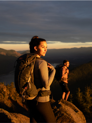 Two hikers with backpacks stand on a rocky outcrop at sunset, overlooking a valley with distant mountains near Suncadia Resort. The breathtaking view captures the essence of Washington's wilderness, while nearby Cle Elum restaurants await with cozy meals after their adventure.