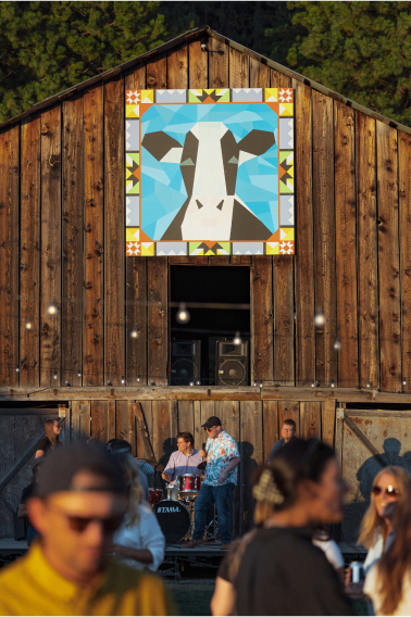 People gather in front of a wooden barn with a geometric cow mural at the Suncadia Resort in Washington. A band plays instruments near the entrance, setting a lively atmosphere that's perfect for exploring nearby Cle Elum restaurants.