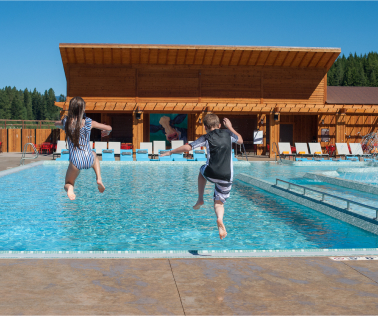 Two children in swimwear jump into an outdoor pool at Suncadia, with a charming wooden building and lounge chairs in the background.