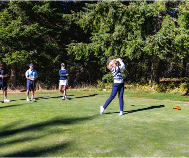 A person swings a golf club on a green at Suncadia Resort, surrounded by three onlookers, with trees in the background.