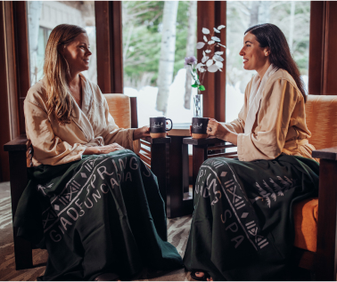 Two people in robes sit on chairs, holding mugs, with spa blankets draped over their laps. A vase with flowers is in the background, evoking the tranquility one might find while exploring Suncadia real estate or unwinding near Cle Elum restaurants.