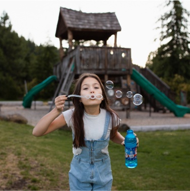 A girl blows bubbles, holding a bottle of solution, her laughter echoing through the air as it mingles with the cheerful ambiance of a wooden playground at Suncadia Resort.