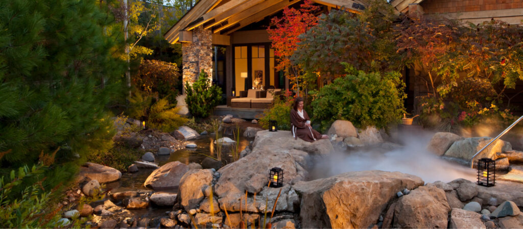 A person relaxes on rocks by a small pond in a garden with lush greenery, reminiscent of the tranquil atmosphere found at Suncadia Resort. In the background, a charming wooden house completes this serene scene.