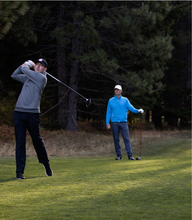 On the lush golf course at Suncadia Resort, one person gracefully swings a golf club, while the other observes amidst the stunning backdrop. Towering trees frame the scene under a clear sky, capturing a serene moment in this picturesque setting.