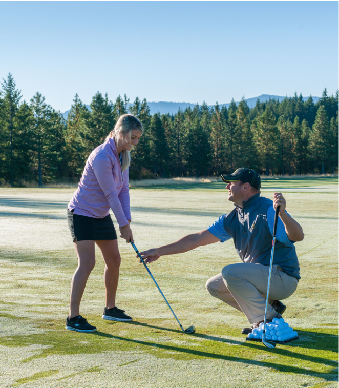 A golf instructor kneels on the lush grass at Suncadia Resort Washington, guiding a woman holding a club. Nearby, practice balls are scattered amidst trees and hills, creating an ideal setting just a stone's throw from charming Cle Elum restaurants.