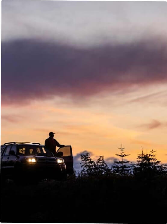 A silhouette of a person stands by a car at dusk, with the colorful sky and trees reflecting the serene beauty of Suncadia.