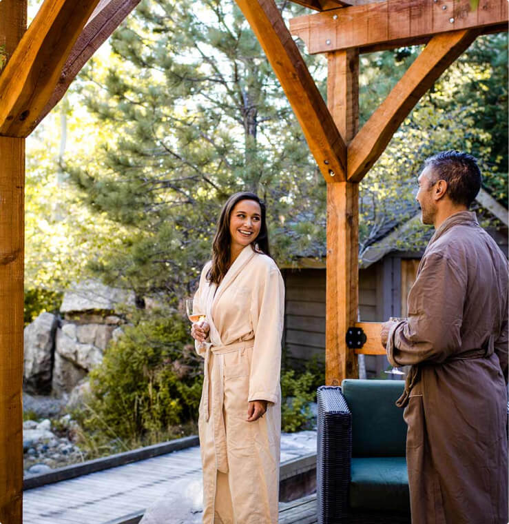 Two people in robes stand outdoors on a wooden deck at Suncadia Resort, surrounded by lush greenery, each holding a drink.