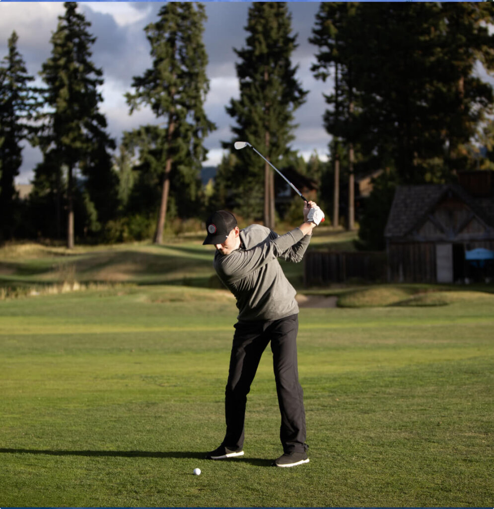 A person in a gray sweater and black pants is skillfully swinging a golf club at the picturesque Suncadia Resort, surrounded by lush trees.