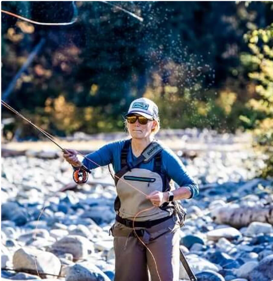 Amidst the tranquil beauty of a rocky riverbed, where lush trees frame the scene, a person in fishing gear and sunglasses enjoys fly-fishing. This picturesque setting captures the serene lifestyle often sought by those exploring Suncadia real estate.