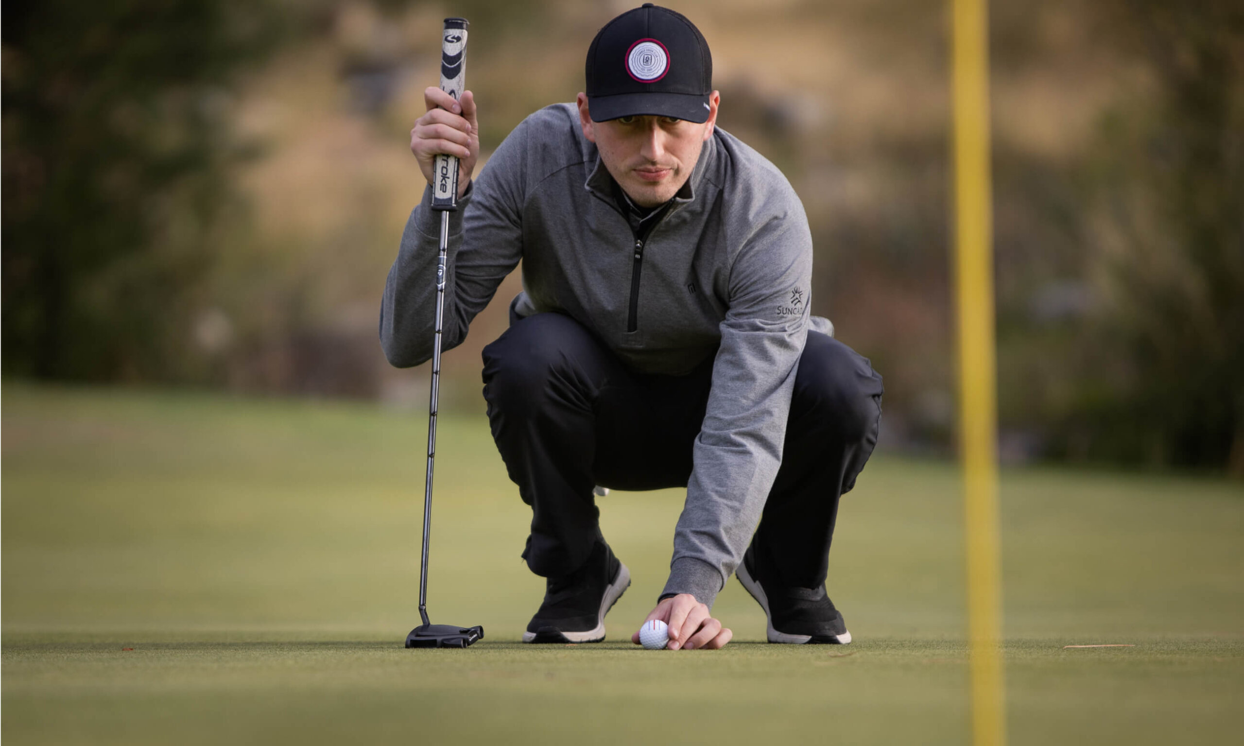 At Suncadia Resort, a person in a gray jacket and black cap crouches on a golf green, skillfully positioning a golf ball next to their putter.